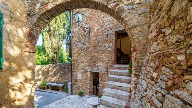 Una escalera de piedra que sube hacia una puerta, rodeada de muros de piedra rústicos y un pequeño banco.