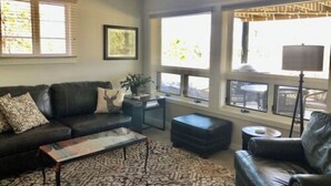 A living room with a black leather sofa, a coffee table, and a patterned rug. There are two windows with blinds, a framed picture on the wall, and a lamp on a side table. - 2 bedroom 1 bath close to downtown and RMNP- sleeps 4-6 (Estes Park)