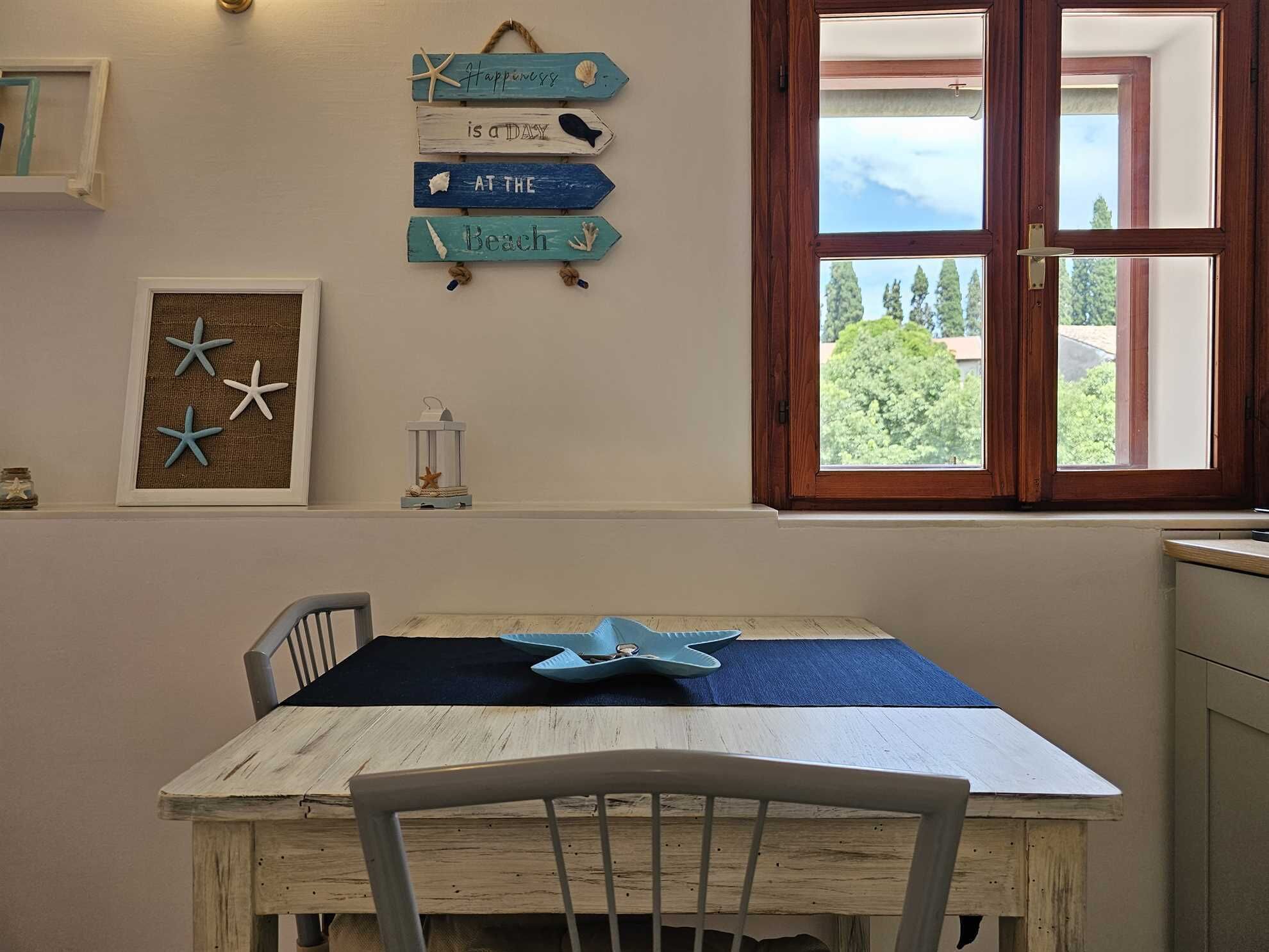Een zithoek met een houten tafel en stoelen, een raam met uitzicht op bomen, en een muur met een bord waarop staat: "Happiness is a DAY AT THE BEACH".
