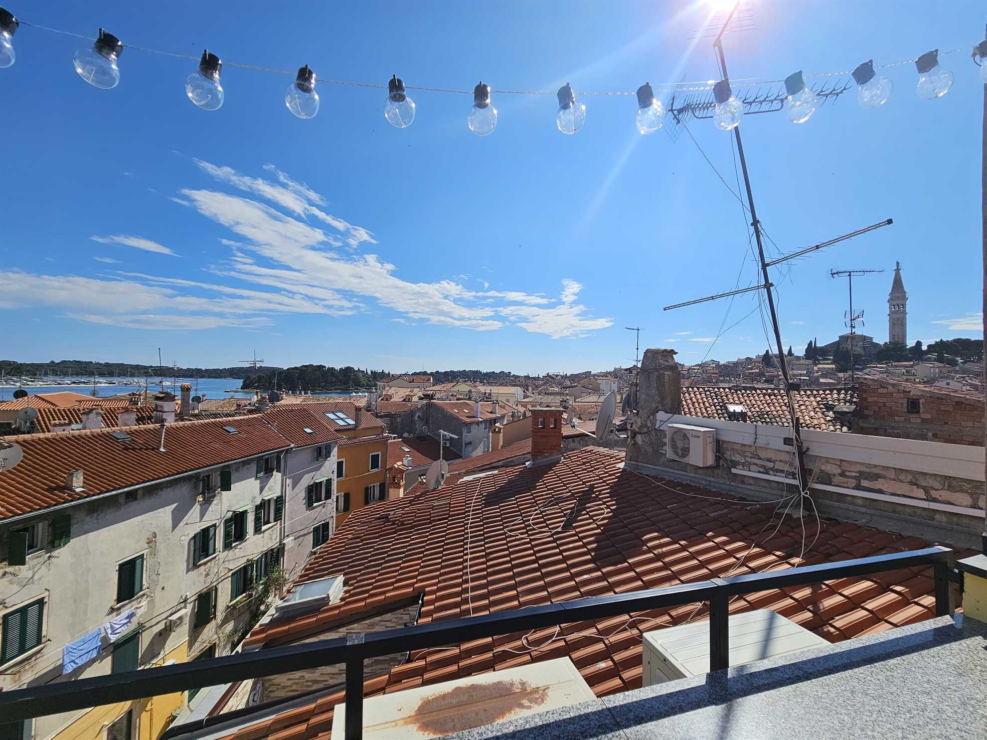 A rooftop terrace with red-tiled roofs, a clear blue sky, and a distant view of a cityscape including a tower.