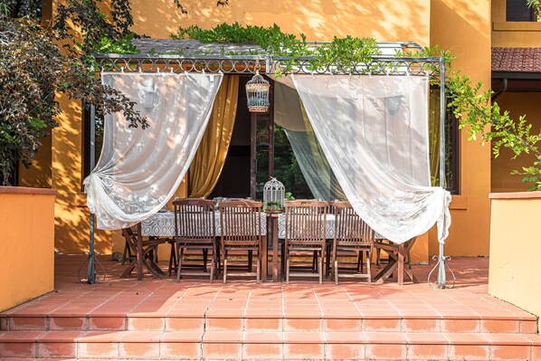 An outdoor patio with a covered table and chairs, surrounded by a yellow wall and greenery.