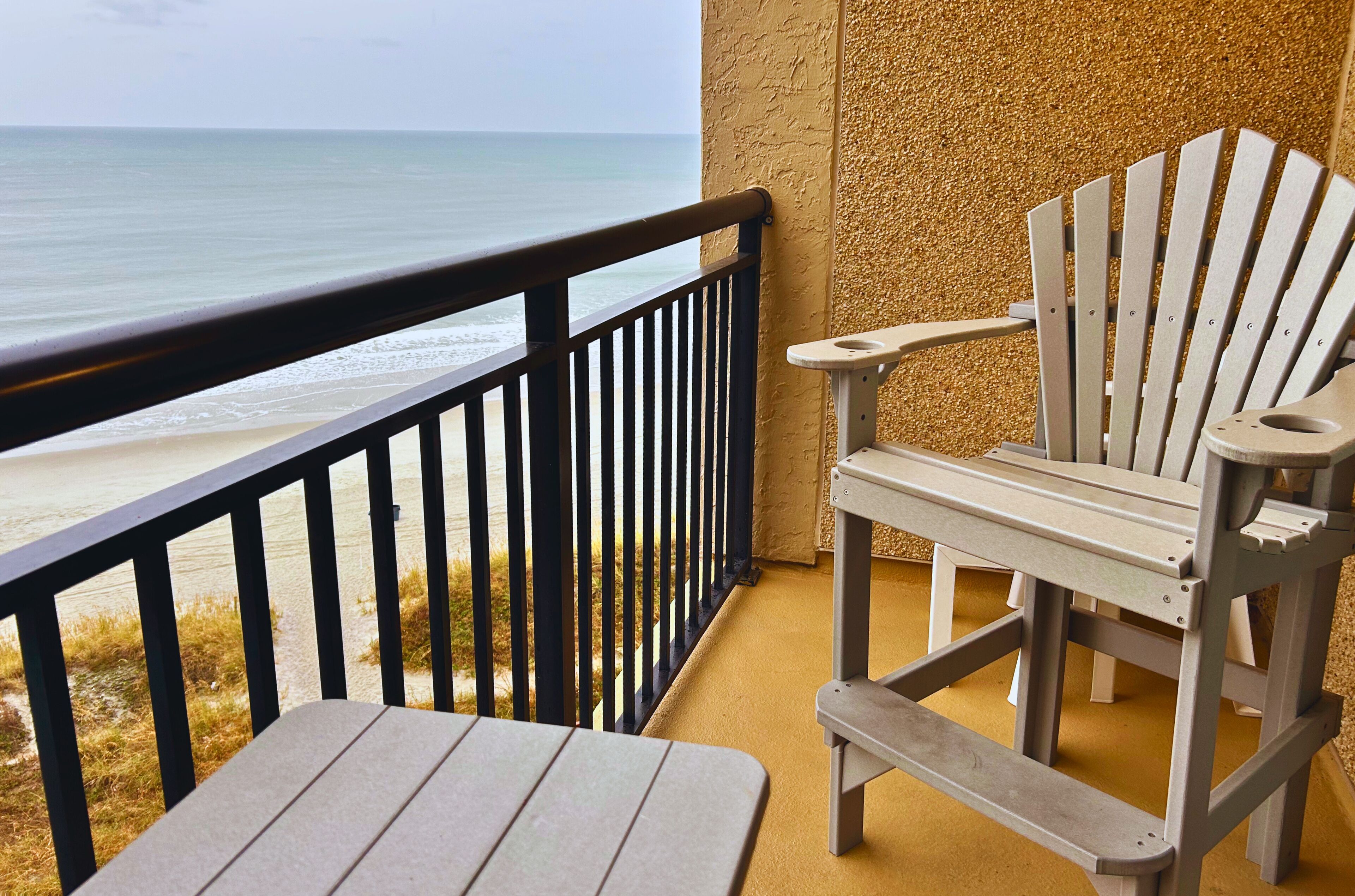 A balcony with a view of the beach, a wooden chair, and a small table.