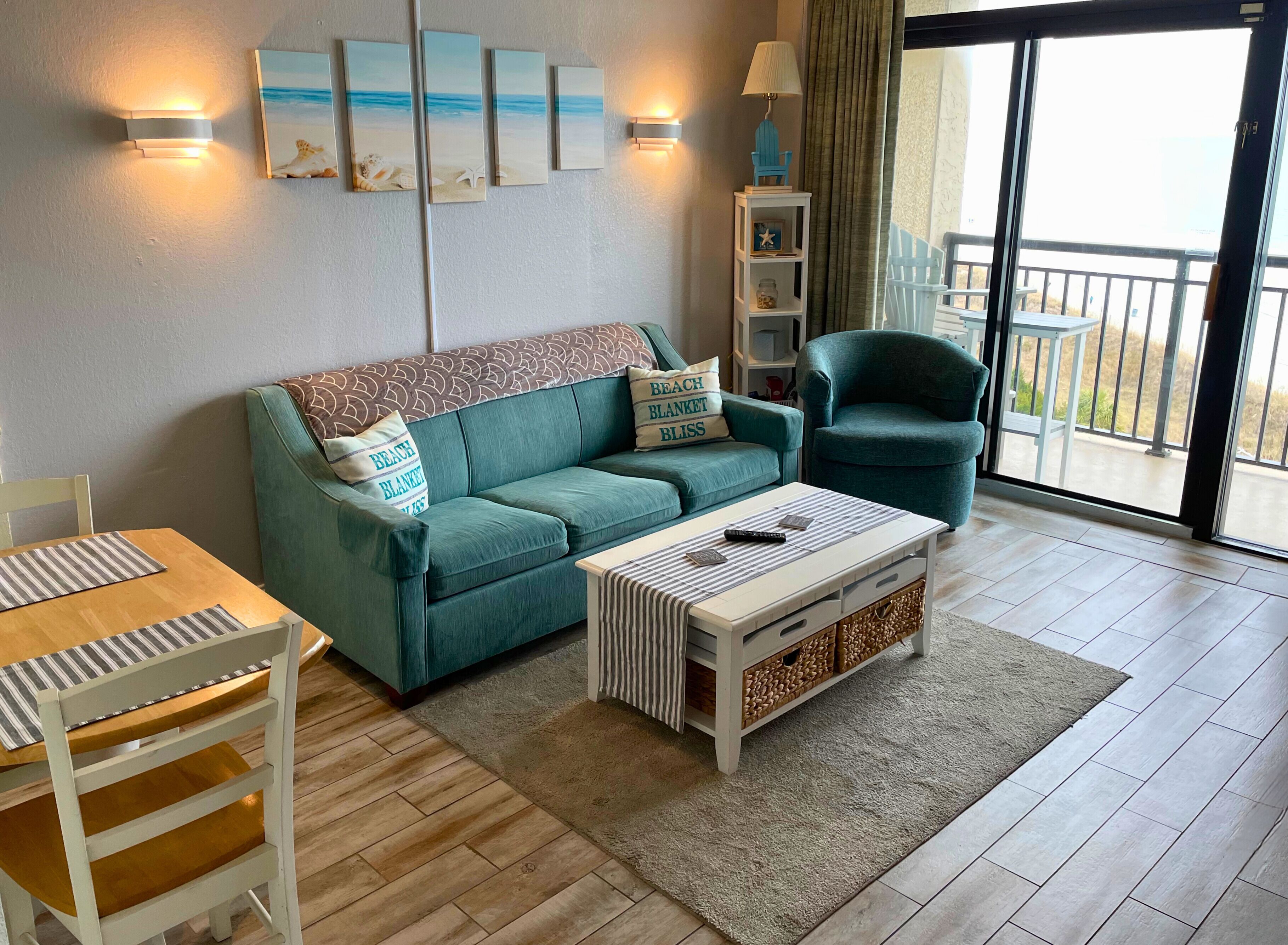 Een woonkamer met een blauwgroene bank, een gestreepte salontafel, een houten stoel en uitzicht op het balkon door een glazen deur.