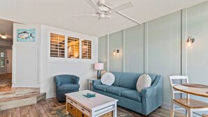 Una sala de estar con un sofá azul, una mesa de centro blanca, un sillón azul y una mesa de comedor de madera.
