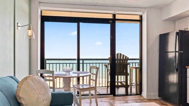 Un balcón con vista al mar, una mesa de comedor con sillas, un sofá y un refrigerador.