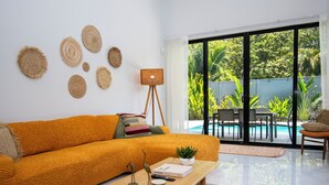 Un salon lumineux avec un grand canapé orange, une table basse en bois et un lampadaire. Les murs sont ornés de tapisseries tissées, et de grandes portes vitrées offrent une vue sur une piscine.