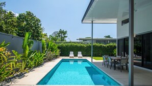 Une piscine extérieure moderne, dotée de chaises longues et d’un espace repas, entourée d’une végétation luxuriante et surplombée par un ciel dégagé.