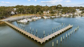 A marina with boats docked, a wooden pier, and houses in the background.