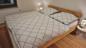 A single bed with a wooden frame, a white and gray checkered bedspread, and two pillows. There is a bedside table with a lamp and a small white object on it.