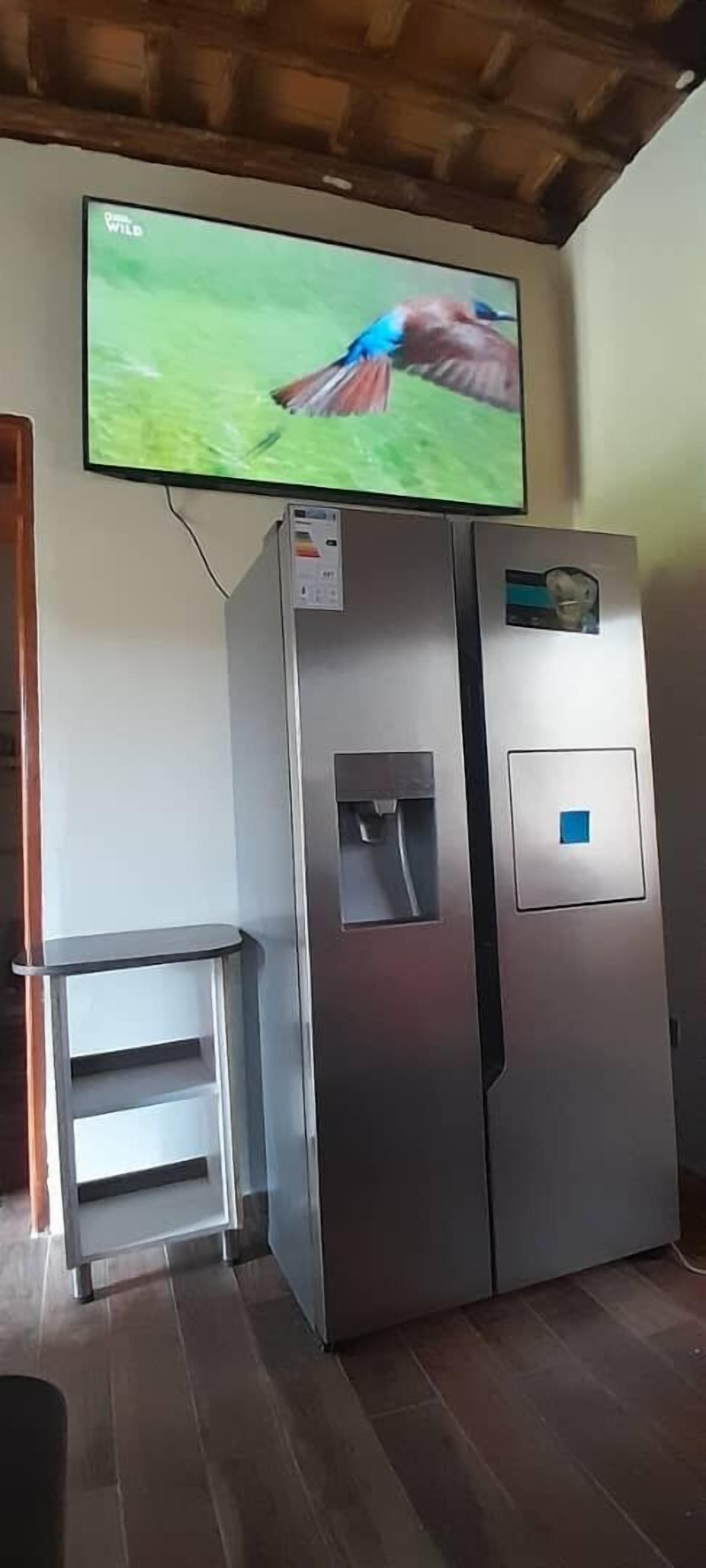 A room with a wall-mounted television displaying a nature scene, a stainless steel refrigerator, and a small white cabinet.