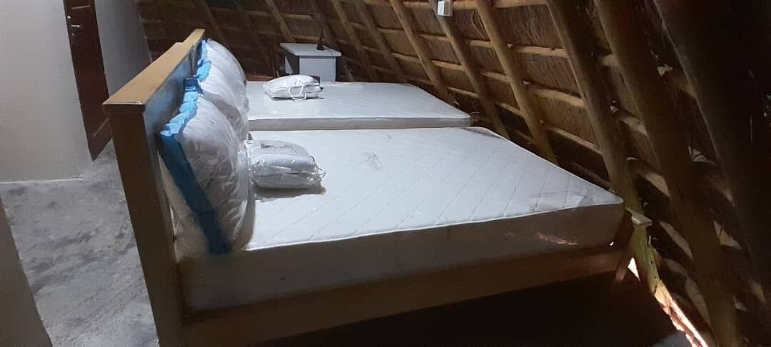 Una habitación con techo inclinado de madera, una cama con colchón y almohadas, y una pequeña mesita de noche.
