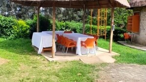 A thatched-roof outdoor dining area with a table and chairs, surrounded by greenery and a water tank in the background. - White Cat Farm Hut Inn (Mashonga)