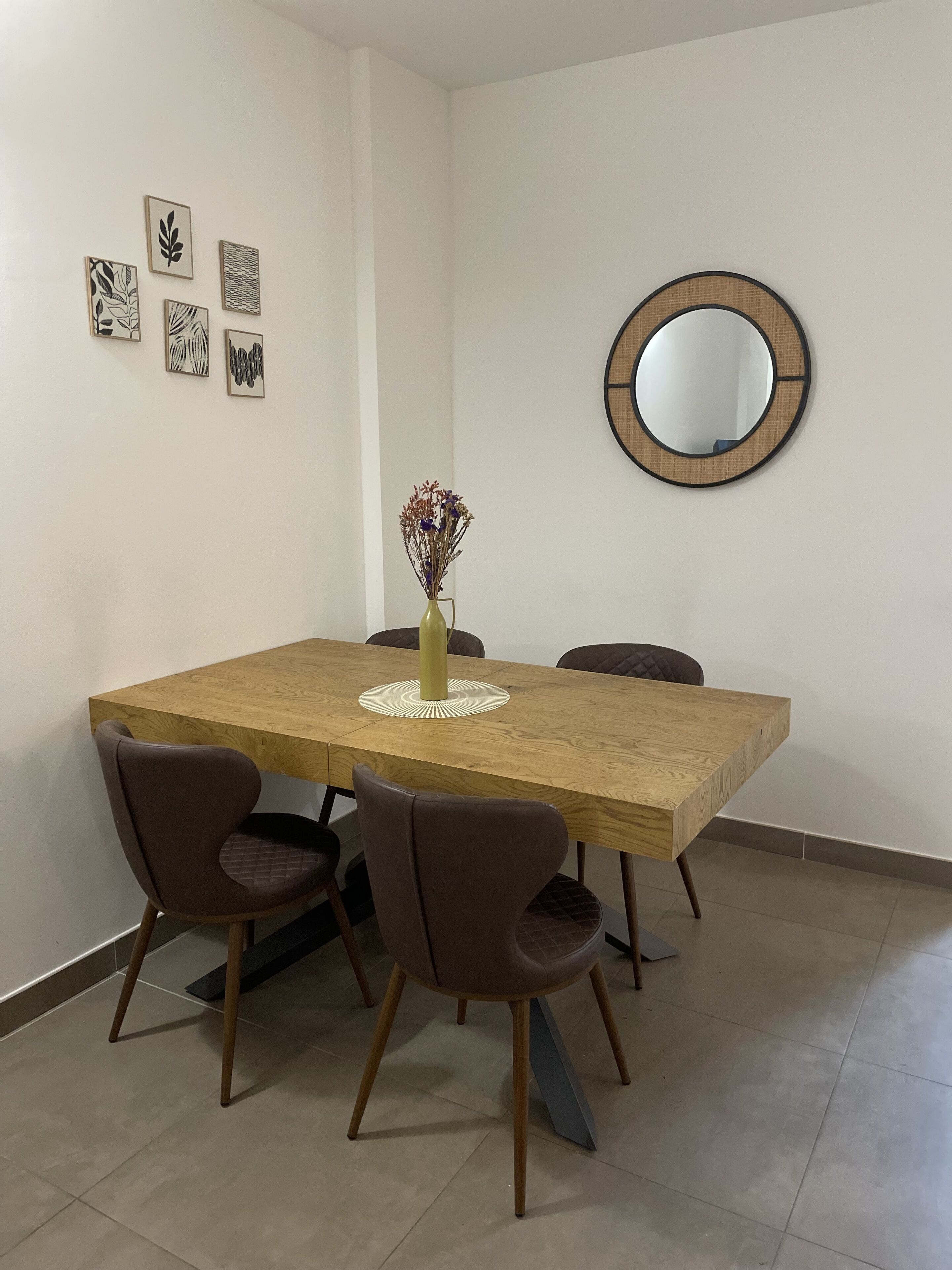 A dining area with a wooden table, six chairs, a vase with dried flowers, and a round mirror on the wall.