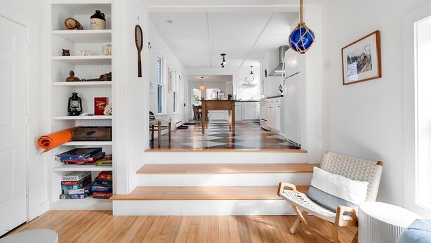 A modern interior with a staircase leading to a dining area, a seating area with a woven chair, and a kitchen visible in the background.