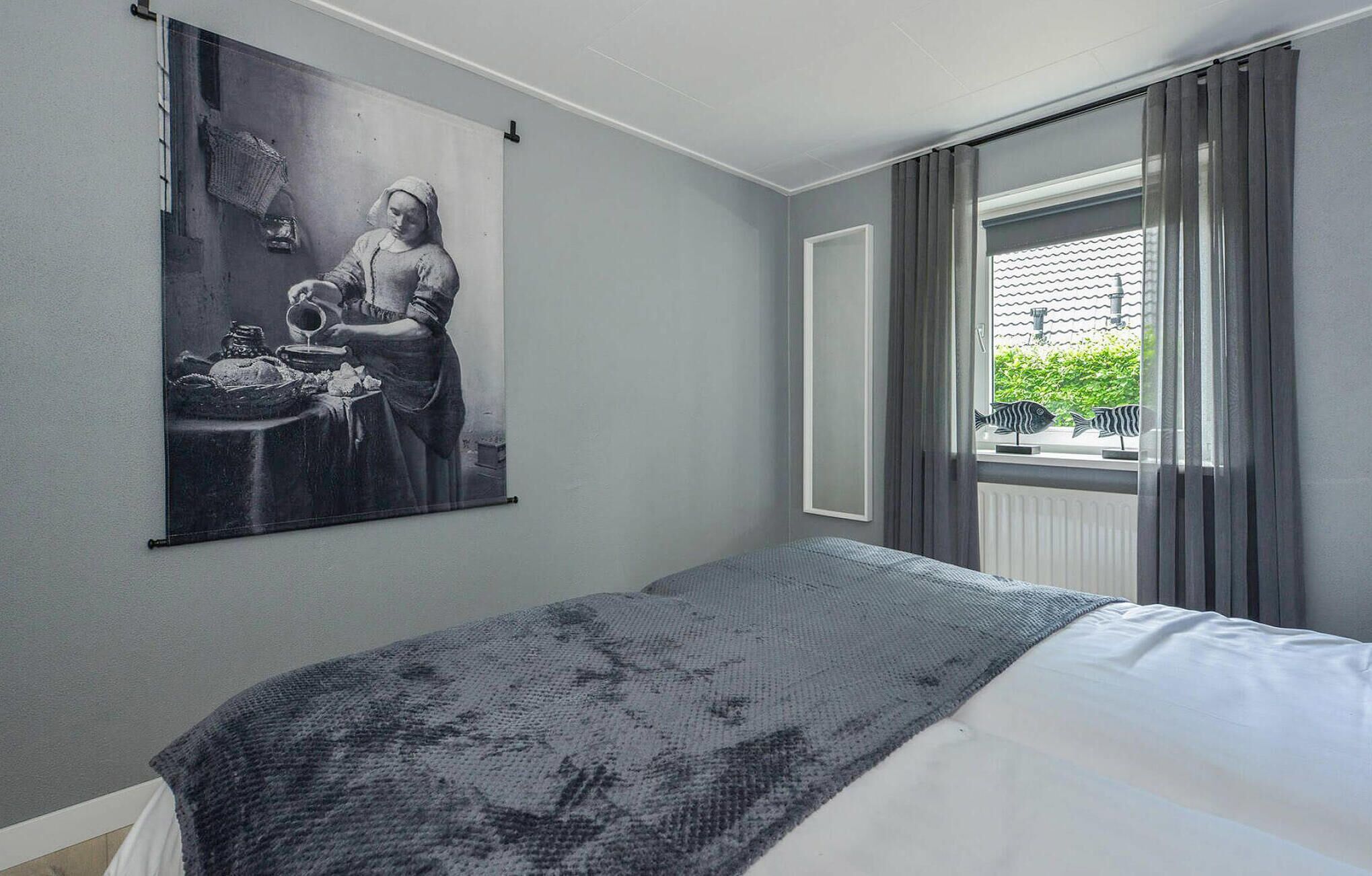 A bedroom with a bed, a large painting, a window with curtains, and a view of greenery outside.