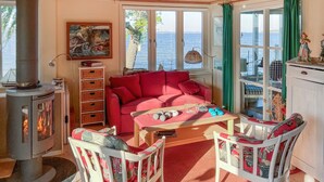 Un salón acogedor con un sofá rojo, una mesa de madera con pesas y vista al mar a través de amplias ventanas.