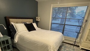 Une chambre à coucher comprenant un lit, une table de chevet, une lampe, un ventilateur de plafond, une fenêtre avec des stores et un tapis à motifs.