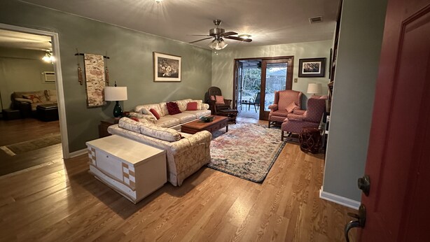 Un salon comprenant un canapé, un fauteuil et un pouf, un ventilateur de plafond et une porte donnant sur un patio.