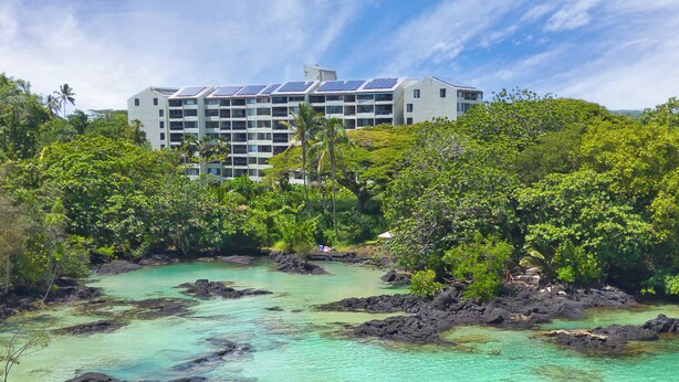 Un hôtel à étages, entouré d’une végétation luxuriante et bordé d’eaux turquoise et cristallines.