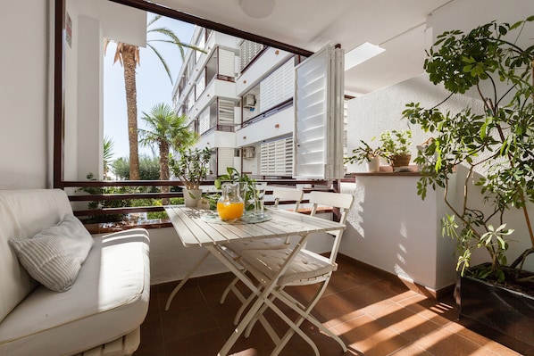 A balcony with a white table, chairs, and a sofa with cushions. There are potted plants and a view of a building with balconies.