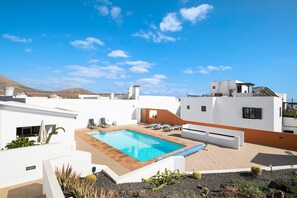 Un espace piscine aménagé sur le toit, avec un ciel d’un bleu éclatant, des bâtiments blancs et une piscine entourée de chaises longues et de plantes.