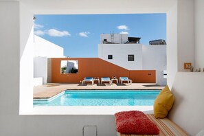 Un espace piscine moderne offrant une vue dégagée sur le ciel, avec une piscine et un coin salon agrémenté d’un coussin rouge.