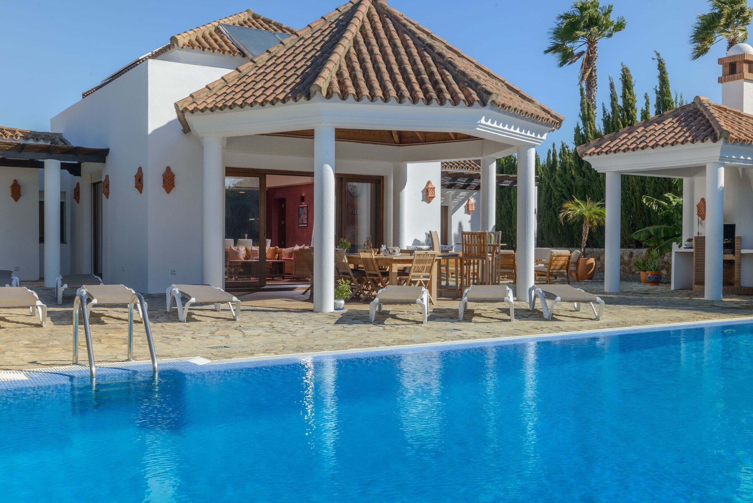 Un espace aménagé au bord de la piscine, avec des chaises longues, un coin salon abrité et un bâtiment doté d’un toit en tuiles.
