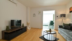 A modern living room with a flat-screen TV on a wooden TV stand, a round coffee table with a rug, a grey sofa, and a dining area visible through an open door.