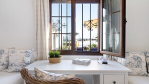 Een kamer met een wit bureau, een rieten stoel en uitzicht op zee door een raam met decoratieve ijzeren tralies.