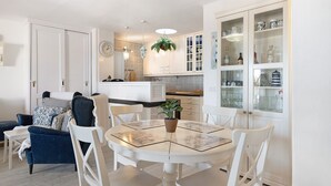 Een eetgedeelte met een ronde tafel, witte stoelen en een potplant. Achtergrond is een keuken met witte kasten en een hangplant.