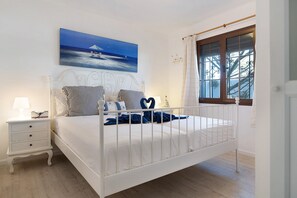 Une chambre avec un lit blanc, une table de chevet, un tableau représentant une scène de plage, une fenêtre avec des rideaux et une vue sur les arbres.