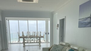 Ett rymligt vardagsrum med en vit lädersoffa, ett soffbord i trä och utsikt över havet genom stora glasdörrar.