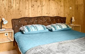 Quarto com cabeceira de madeira, duas mesas de cabeceira com luminárias, e cama com almofadas listradas e colcha azul.