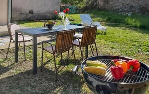 Uma área de refeições ao ar livre com mesa e cadeiras, uma churrasqueira com legumes e um vaso com flores.