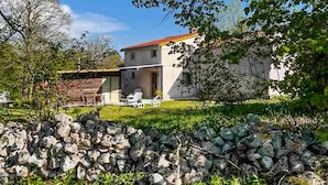 Una casa de dos pisos con un muro de piedra al frente, un patio con mobiliario exterior y un cielo azul despejado.