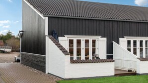 Il s’agit d’un bâtiment à l’extérieur sombre, avec des murs extérieurs blancs et un toit recouvert de tuiles. On y trouve deux portes d’entrée blanches, dotées de fenêtres, ainsi qu’une petite terrasse en bois.
