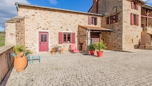 Un edificio de piedra con puerta roja y ventanas, un patio empavedado y mobiliario exterior.