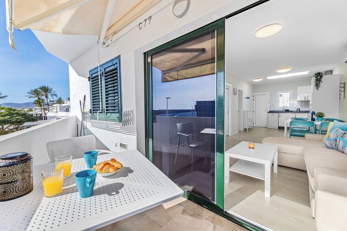 A modern balcony with a dining table, chairs, and breakfast items. Inside, there is a living area with a sofa and a kitchen with white cabinets.