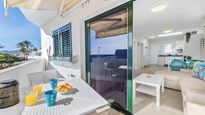 A modern balcony with a dining table, chairs, and breakfast items. Inside, there is a living area with a sofa and a kitchen with white cabinets.