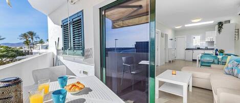 A modern balcony with a dining table, chairs, and breakfast items. Inside, there is a living area with a sofa and a kitchen with white cabinets.