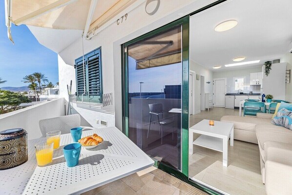 A modern balcony with a dining table, chairs, and breakfast items. Inside, there is a living area with a sofa and a kitchen with white cabinets.