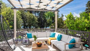 A covered outdoor seating area with a white sofa and cushions, a wooden table with a fruit bowl, and a hanging chair.