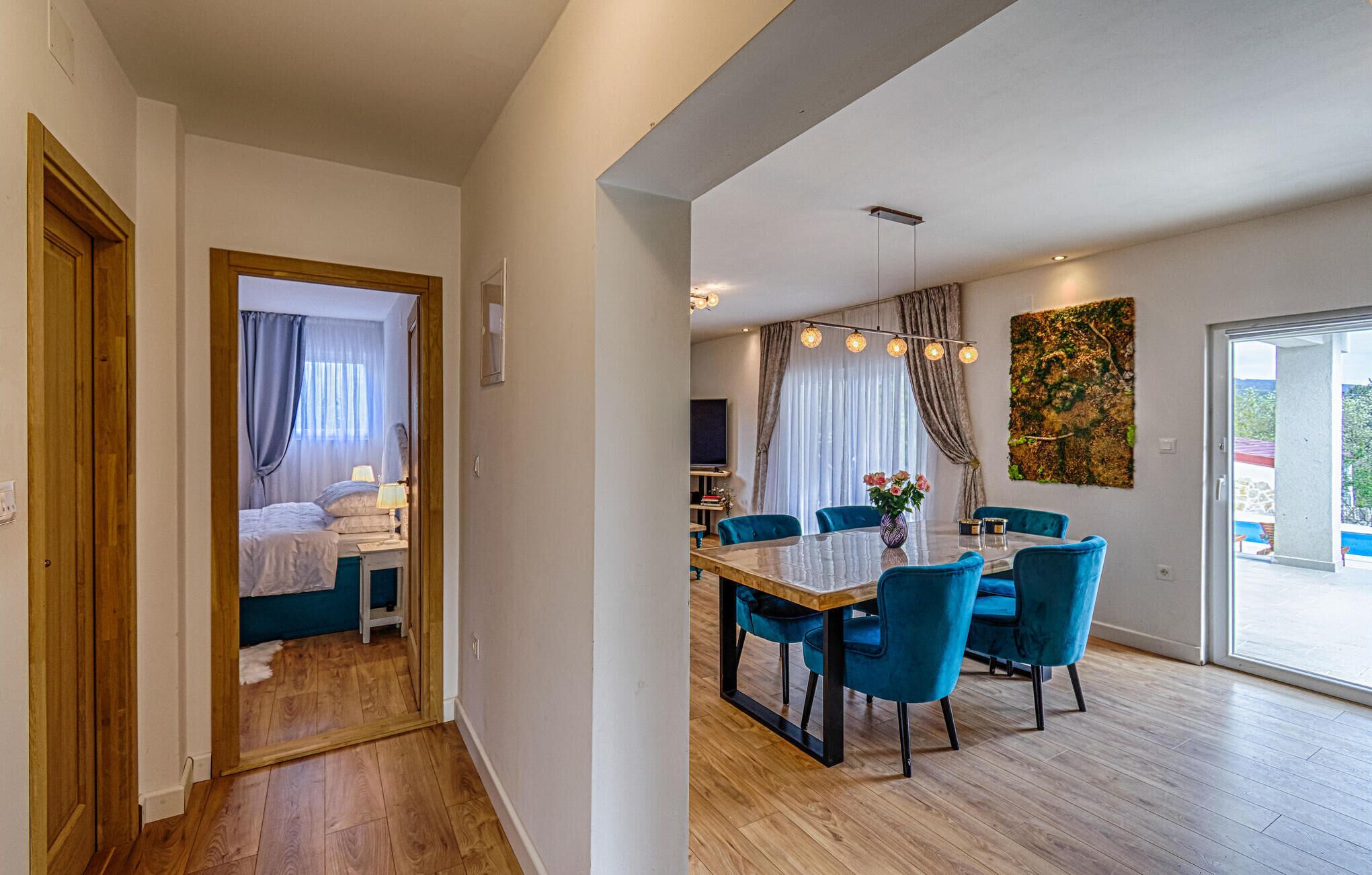 A modern hotel room with a wooden floor, a bed with curtains, a dining area with blue chairs, and a large window with a view.