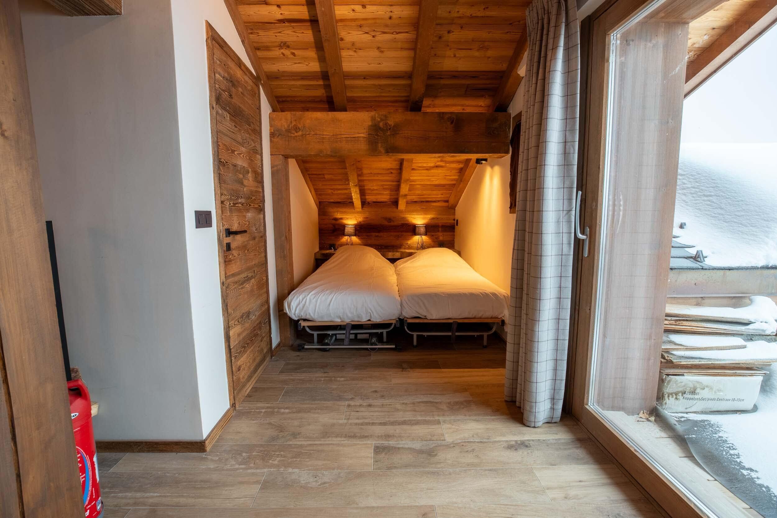 A narrow hallway leading to a room with two beds, wooden ceiling beams, and a window with curtains.