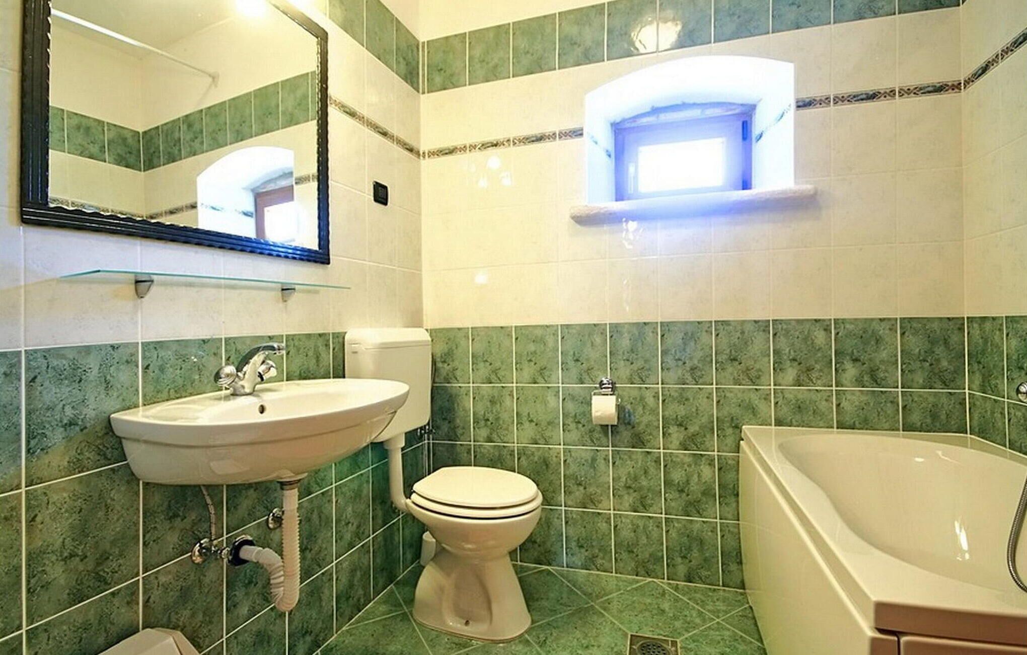 Et badeværelse med vask, toilet og badekar, med grønne fliser og et spejl.