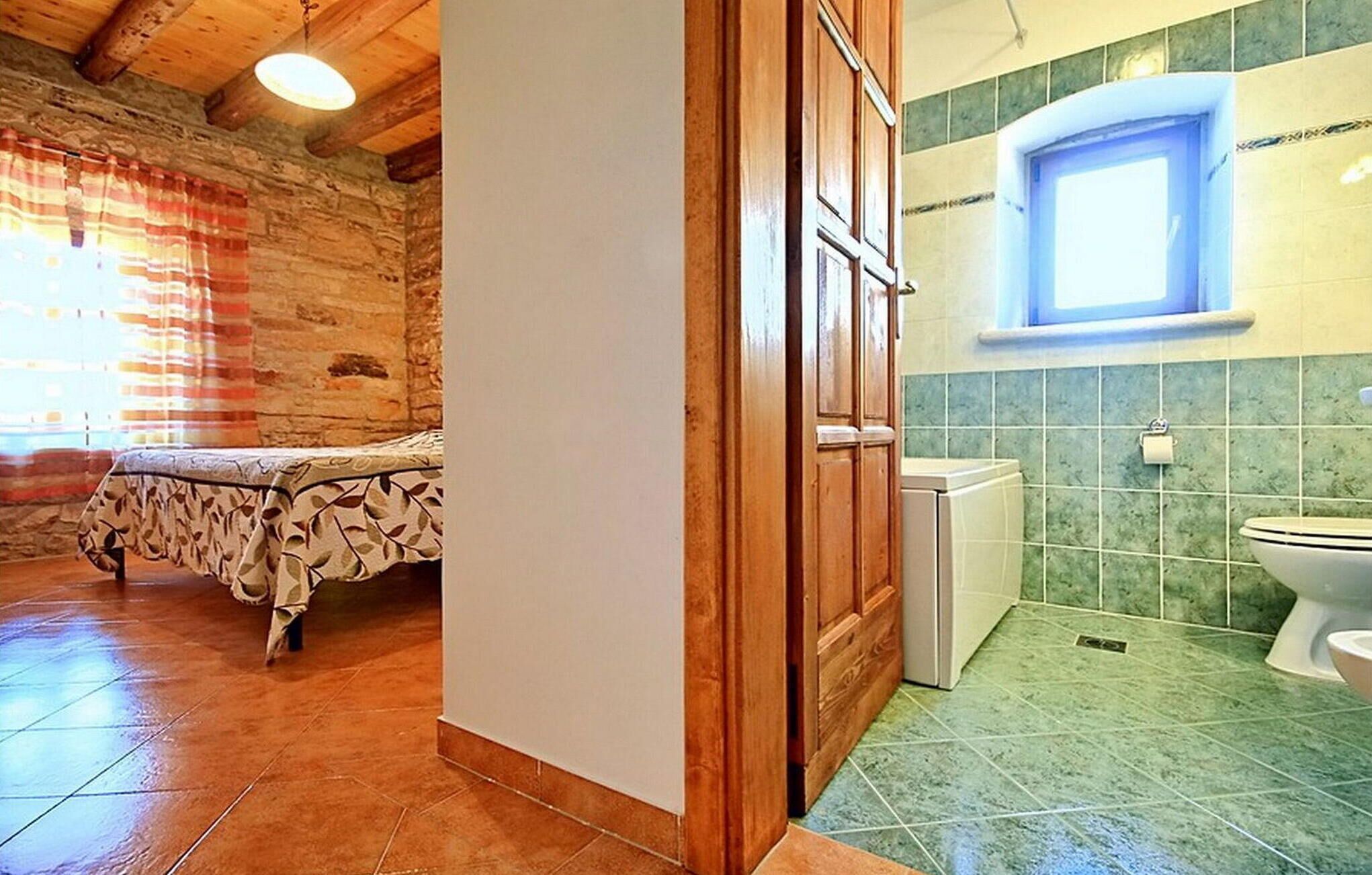 Una habitación con una cama, una puerta de madera, un ventanal pequeño y un baño con inodoro y lavabo.