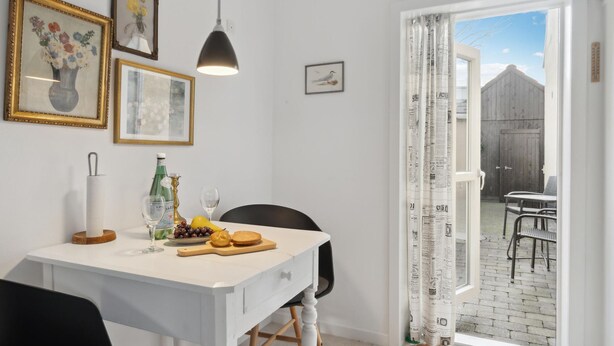 Een eetgedeelte met een tafel gedekt voor twee personen, met een fles, glazen en fruit. Er hangen afbeeldingen in lijsten aan de muur en een hangende hanglamp.