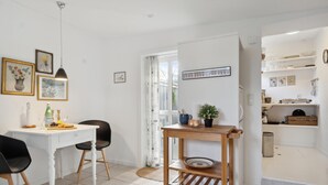 Een moderne keuken met witte kasten, een houten tafel en een zwarte stoel. Er hangen afbeeldingen in lijsten aan de muur, een raam met gordijnen en een plank met keukengerei.