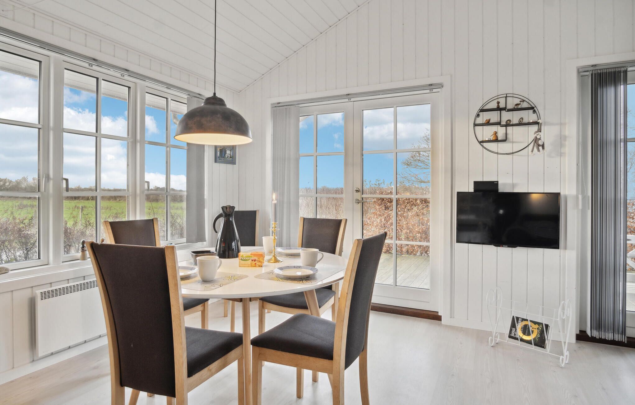A dining area with a table and chairs, a pendant light, a television mounted on the wall, and large windows with a view of the outdoors.