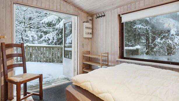 A wooden cabin room with a bed, a chair, a window, and a door leading to a balcony with a view of snow-covered trees. - Stunning home in Silkeborg with WiFi (Silkeborg)